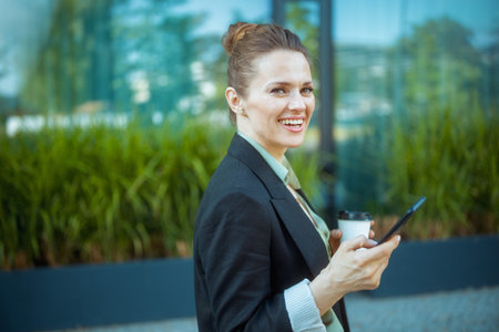 smiling modern 40 years old business woman in business district in black jacket with smartphone and cup of coffee.の写真素材