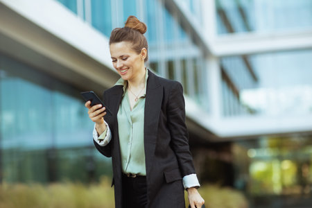 happy modern middle aged woman worker near office building in black jacket using smartphone and walking.の写真素材