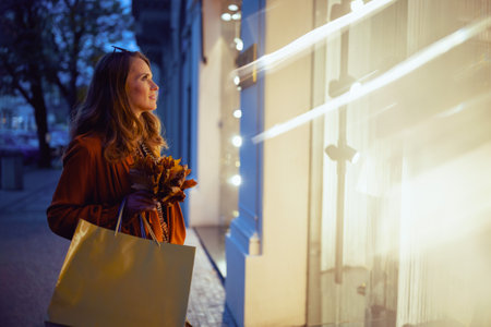 40 years old woman in brown trench coat with shopping bags and autumn yellow leaves near store in the city.の写真素材