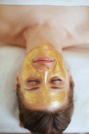 Healthcare time. Upper view of relaxed modern middle aged woman in spa salon with golden cosmetic mask on face laying on massage table.の写真素材
