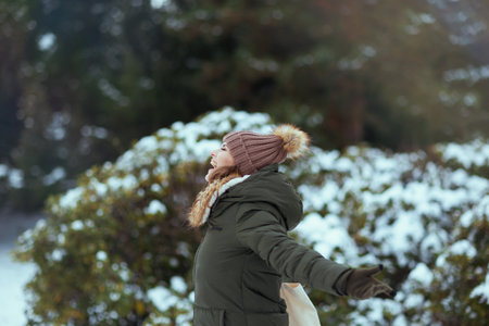 happy modern woman in green coat and brown hat outdoors in the city park in winter with beanie hat near snowy branches.の写真素材