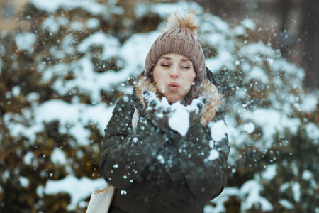 modern 40 years old woman in green coat and brown hat outdoors in the city park in winter with mittens and beanie hat blowing snow near snowy branches.の写真素材