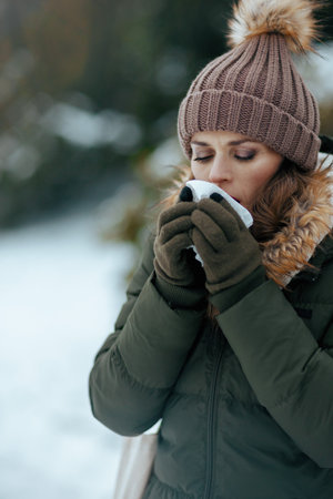 sad modern middle aged woman in green coat and brown hat outdoors in the city park in winter with mittens, napkin blowing nose and beanie hat.の写真素材