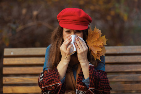Hello autumn. stylish middle aged woman in red hat with autumn leaves, scarf and napkin blowing nose while sitting on bench in the city park.の写真素材