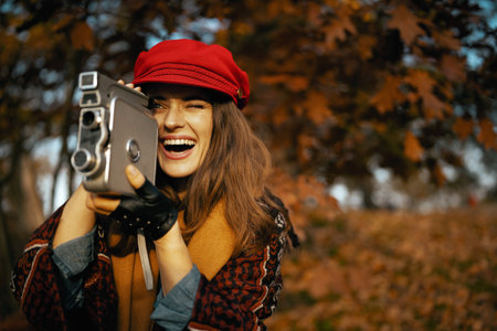 Hello autumn. smiling stylish woman in red hat with retro video camera, scarf and gloves in the city park.の写真素材