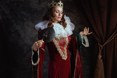 medieval queen in red dress with fan, white collar and crown on dark gray background.