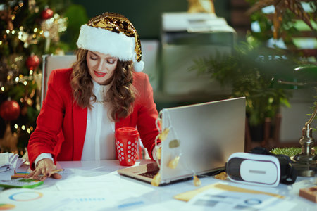 Christmas time. happy elegant 40 years old small business owner woman in santa hat and red jacket with documents and laptop working in modern green office with Christmas tree.の写真素材