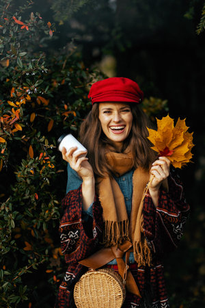 Portrait of happy modern woman in red hat with autumn leaves, scarf, bag and coffee in the city...