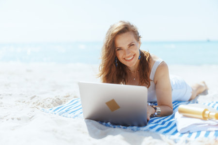 happy stylish middle aged woman on the beach with straw bag, laptop and striped towel.の写真素材