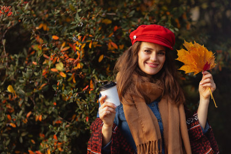 Hello autumn. smiling young woman in red hat with autumn leaves, scarf and coffee in the city park.の写真素材
