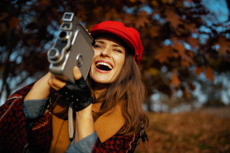 smiling stylish middle aged woman in red hat with retro video camera, scarf and gloves in the city...