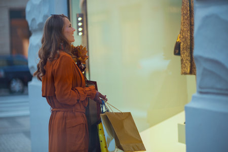 Hello autumn. smiling trendy woman in brown trench coat with shopping bags and autumn yellow leaves near store in the city.の写真素材