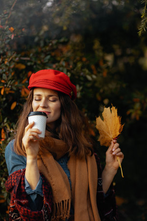 Hello autumn. modern 40 years old woman in red hat with autumn leaves, scarf and coffee in the city park.の写真素材