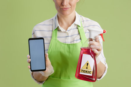 Spring cleaning. Closeup on confused modern female in green apron and rubber gloves isolated on green background with bottle of toxic cleaning product showing smartphone screen.の写真素材