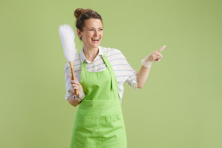 Spring cleaning. smiling modern woman in green apron and rubber gloves on green background with dust broom.の写真素材