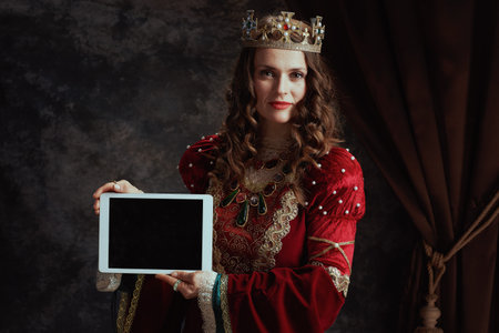 medieval queen in red dress with crown showing tablet PC blank screen on dark gray background.の写真素材