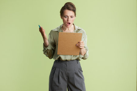 A woman looks shocked while holding a clipboard, her mouth agape. Dressed professionally in green and grey, she seems taken aback by news.の写真素材