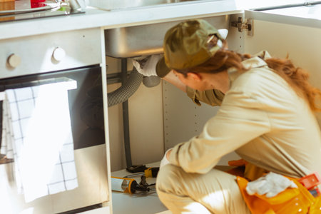 A woman plumber kneels under a kitchen sink, inspecting pipes for repair. The scene highlights focused home maintenance, DIY, and skilled problem-solving.の写真素材