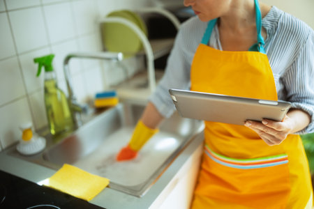 Woman in striped shirt and yellow apron uses a tablet while cleaning a stainless steel sink, showing modern multitasking in a bright kitchen.の写真素材