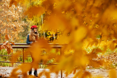 Hello autumn. happy stylish woman in jeans shirt and red hat with autumn leaf and scarf walking in...
