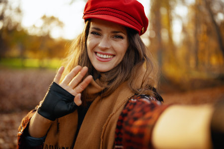 Hello autumn. smiling elegant woman in red hat with scarf and gloves talking video call in the city park.の写真素材