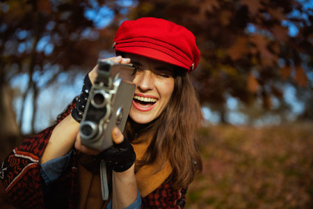 Hello autumn. smiling elegant female in red hat with retro video camera, scarf and gloves in the city park.の写真素材