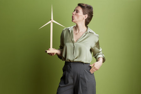 A woman affectionately looks at a wooden wind turbine model, symbolizing support for environmental awareness and sustainable energy against a solid green background.の写真素材