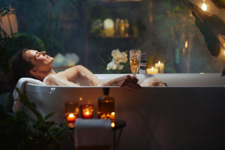A middle-aged woman with her hair pulled back relaxes in a bubbly bathtub, holding a champagne glass. Lit candles, lush greenery, and soft, warm lighting create a luxurious, spa-like atmosphere for self-care and indulgence.の写真素材