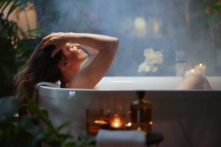A serene woman with long brown hair relaxes in a steaming white bathtub, eyes closed, surrounded by soft candlelight and lush green plants. This tranquil scene evokes a sense of self-care, peace, and spa-like luxury.の写真素材