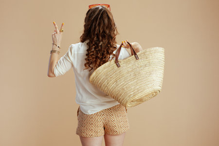 Beach vacation. Seen from behind woman in white blouse and shorts isolated on beige with straw bag and sunglasses showing victory gesture.の写真素材