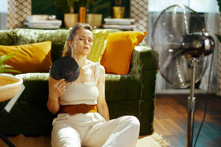 A woman in her 30s-40s, eyes closed and head tilted back, battles the summer heat from her sunlit living room. She uses a small hand fan while a larger standing fan provides additional cooling relief.の写真素材
