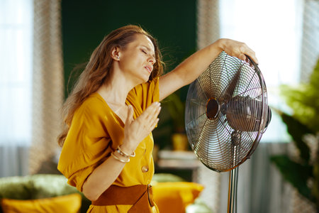 A woman with grimacing expression & visible perspiration struggles with intense summer heat indoors. She seeks relief, fanning herself while leaning on a standing fan, conveying overheating discomfortの写真素材