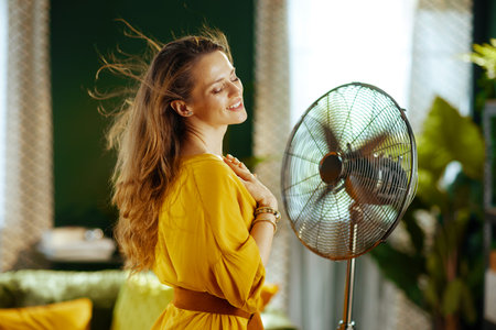 An adult woman finds serene bliss in her living room, as she enjoys the cool breeze from a rotating metal floor fan. Her hands rest on her chest, conveying profound relief from summer heatの写真素材