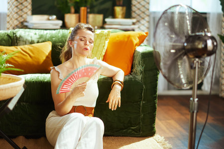 A woman with closed eyes sits on a wooden floor, leaning against a green couch, fanning herself with a hand fan. A standing fan provides additional aid in her bright living room, combating summer heatの写真素材