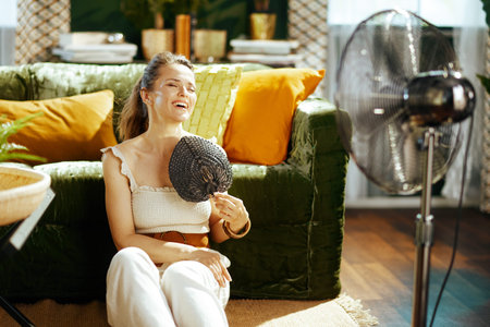 A smiling woman with eyes closed enjoys refreshing breeze from large floor fan & hand fan in bright...