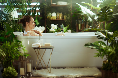 A woman with her hair in a bun enjoys a peaceful bubble bath, eyes closed, bathed in warm sunlight, surrounded by lush green plants and spa products in a tranquil bathroom.の写真素材