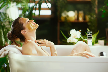 A serene side profile of a woman with a yellow face mask relaxes with eyes closed in a bubble bath....