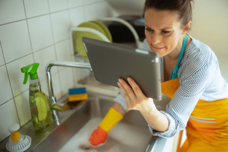 A woman in gloves and apron uses a tablet while facing a clogged kitchen sink, searching for help or instructions to resolve a drain issue.の写真素材