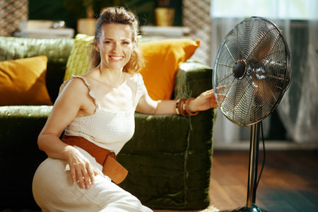 A smiling young woman kneels in her living room beside a powerful metallic standing fan. Captures...