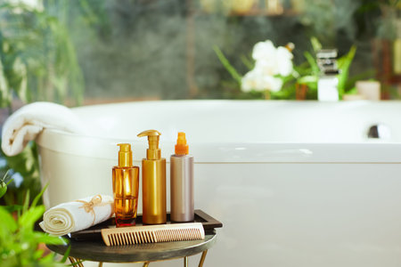 Hair & bath products, bottles, rolled towel, & wooden comb, arranged on a side table. White bathtub...