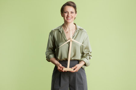A smiling woman in a green blouse and gray pants holds a wooden wind turbine model against a light green backdrop, symbolizing optimism for sustainable energy.の写真素材
