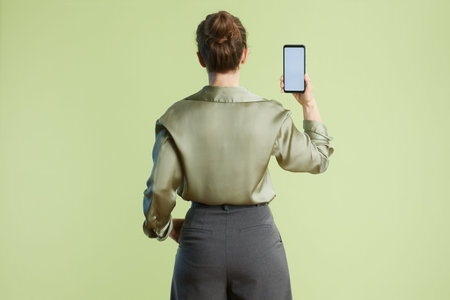 A woman stands with her back to the viewer, holding a blank smartphone. The green blouse and grey...