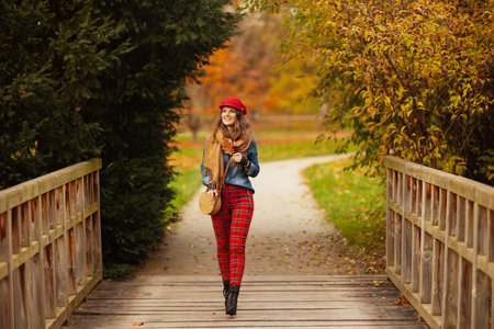 Hello autumn. Full length portrait of smiling modern woman in jeans shirt and red hat with autumn leaf, scarf and bag walking in the city park.の写真素材