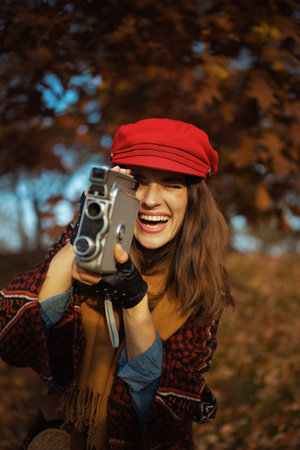 Hello autumn. happy trendy 40 years old woman in red hat with retro video camera, scarf and gloves in the city park.の写真素材