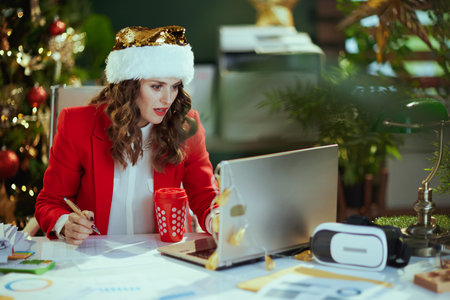Christmas time. stylish small business owner woman in santa hat and red jacket with documents, cup of hot chocolate and laptop using video chat and working in modern green office with Christmas tree.の写真素材
