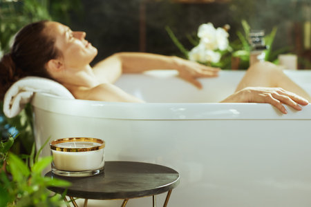 Serene side view of a relaxed woman with brown hair in a white bathtub, head on towel, hands on tubs...