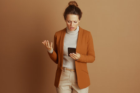 A businesswoman with a frustrated and confused expression gestures while looking at her smartphone. The image captures the annoyance of technological issues or a digital communication challenge.の写真素材