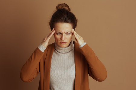 An adult woman with pained expression and furrowed brow presses both hands to her temples, conveying discomfort or severe headache. Dressed in beige turtleneck, she battles stress or painの写真素材