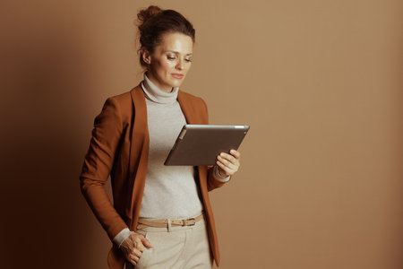 A professional woman in a brown blazer and beige turtleneck intently uses a digital tablet. Her focused expression against a plain background conveys concentration and modern productivity.の写真素材