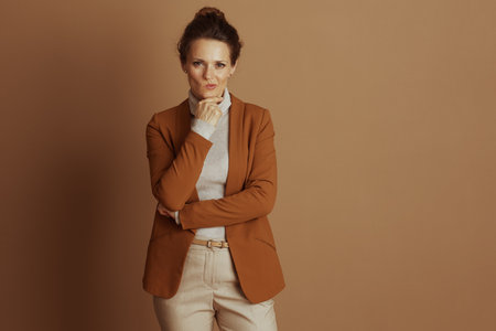 A thoughtful businesswoman, hand on chin, gazes at camera, pensive against a plain brown background. Her beige turtleneck and open rust-colored blazer suggest contemplation or questioning.の写真素材